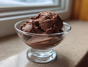 A scoop of rich, airy chocolate mousse served in a clear glass dish near a window.