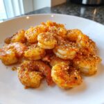 A close-up of seasoned, cooked air fryer frozen shrimp piled on a white plate, sprinkled with spices.