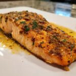 Close-up of a beautifully seasoned and cooked piece of air fryer salmon resting in butter sauce on a white plate.