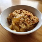 A close-up of a warm serving of amazing apple crumble with golden topping in a white bowl.
