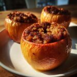 Three golden-brown baked apples filled with a spiced, sugary crumble, sitting on a white plate.