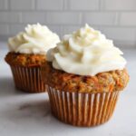 Two moist carrot cake muffins topped with thick swirls of white cream cheese frosting.
