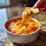 A hand dips a tortilla chip into a hot, bubbly bowl of chili cheese dip, showing a long, satisfying cheese pull.