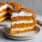 A tall slice of amazing pumpkin crunch cake showing three layers of orange cake, white cream filling, and a crumb topping.