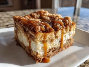 Close-up of a thick slice of apple cheesecake bars with a crumb topping and rich caramel sauce drizzled over it.