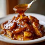 Close-up of thick, syrupy apple pie filling recipe being spooned over a base, showing rich cinnamon color.