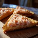 Close-up of three golden baked shrimp toast triangles topped with pink shrimp paste on a wooden cutting board.