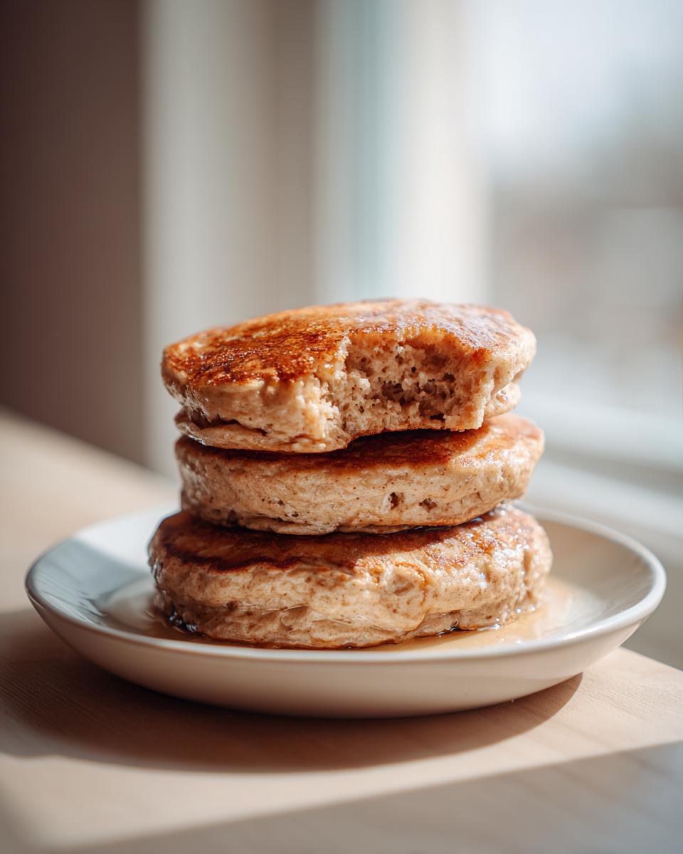 A stack of three fluffy banana oatmeal pancakes drizzled with syrup, with a bite taken out of the top one.