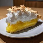 A close-up slice of rich lemon meringue pie showing bright yellow filling and toasted meringue peaks.