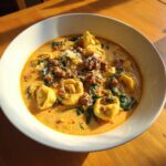 A close-up of a bowl of creamy tortellini soup topped with crumbled sausage, wilted spinach, and grated Parmesan cheese.