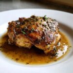 Close-up of a perfectly cooked braised chicken thighs piece sitting in rich pan sauce, topped with herbs.