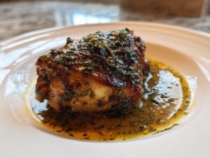 Close-up of a single, perfectly braised chicken thigh coated in a rich, dark glaze and herbs, resting in its sauce.