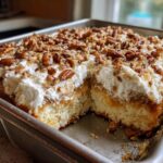 A square slice removed from a rich butter pecan praline poke cake topped with whipped cream and pecans.