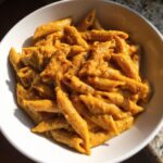 A white bowl filled with penne pasta coated in a thick, vibrant orange butternut squash pasta sauce, seasoned with herbs.