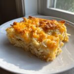 A square serving of cheesy potato bake, featuring shredded potatoes and a melted, golden brown cheddar cheese topping.