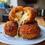 Close-up of crispy fried rice balls stacked, with one broken open showing melted, gooey cheese stretching out.