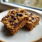 Two chewy banana oatmeal bars topped with visible oats and melted chocolate chips resting on a light plate.