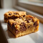 Two thick, chewy chocolate chip cake mix cookie bars sitting on a white surface in bright sunlight.
