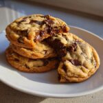 A stack of three chewy chocolate chip cookies, with the top one broken open revealing gooey, melted chocolate.