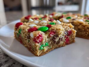 A thick, chewy slice of Christmas cookie bars topped with red, green, and white M&Ms sits on a white plate.