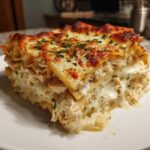 A thick, cheesy slice of chicken alfredo lasagna showing layers of pasta, creamy filling, and a browned cheese topping garnished with parsley.