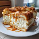 A thick slice of churro cheesecake featuring a creamy white filling, topped with churro pieces, cinnamon sugar, and caramel sauce.