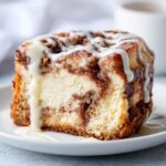 Close-up of a rich slice of cinnamon roll cheesecake topped with white icing drizzle.