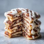 A tall stack of fluffy cinnamon roll pancakes, cut to show the swirl inside, dripping with white cream cheese icing.
