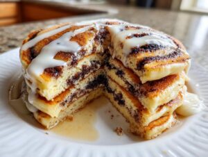 A tall stack of fluffy cinnamon roll pancakes cut open to show the swirl filling, topped with white icing.