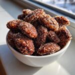 A white bowl filled with glistening, crunchy cinnamon sugar pecans, highlighted by bright sunlight near a window.