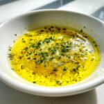 Close-up of melted, bright yellow cowboy butter dipping sauce seasoned with green herbs.