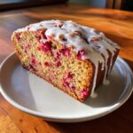 A thick slice of moist cranberry orange bread topped with a thick white glaze, sitting on a white plate.