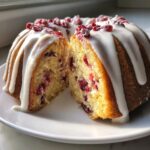 A slice cut from a bundt-style cranberry orange cake, drizzled with white icing and topped with sugared cranberries.