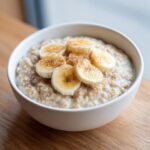 Close-up of creamy banana oatmeal in a white bowl, topped with sliced bananas and a sprinkle of cinnamon.