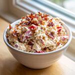 A white bowl filled with creamy blt chicken salad, featuring chunks of chicken, red tomatoes, and topped generously with crispy bacon.