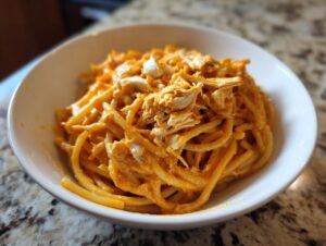 A close-up of creamy buffalo chicken pasta tossed in an orange sauce, topped with shredded chicken, served in a white bowl.