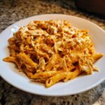 A close-up of creamy, orange-sauced buffalo chicken pasta with shredded chicken served on a white plate.
