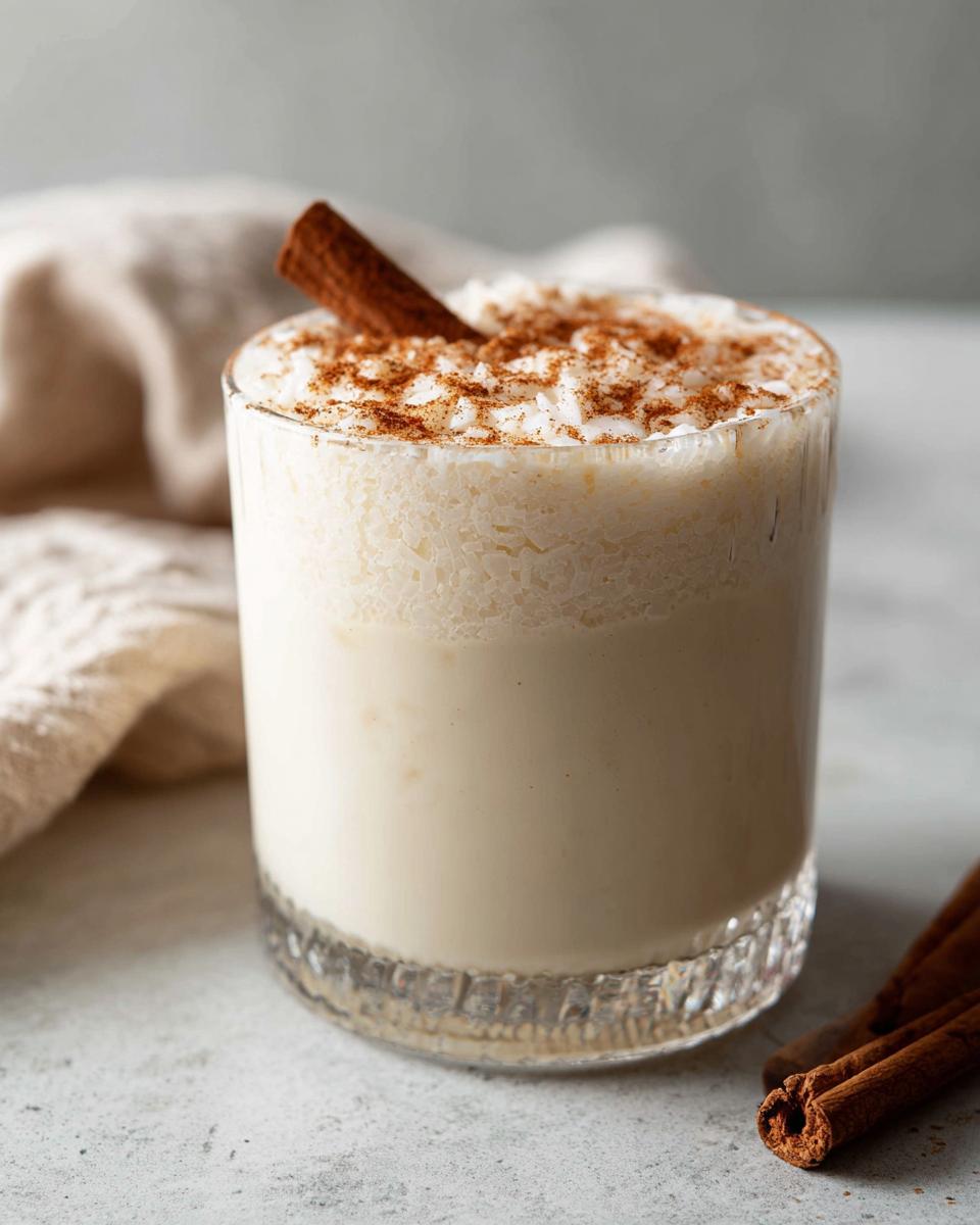 A glass of creamy, frothy coquito topped with ground cinnamon and a whole cinnamon stick.