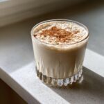 A glass of creamy, light-colored coquito topped generously with ground cinnamon, sitting on a white counter.