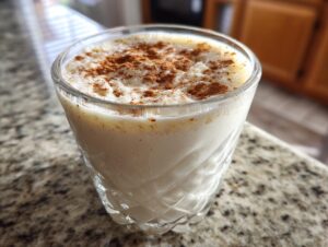 A close-up of a glass filled with creamy homemade coquito, dusted generously with ground cinnamon on top.