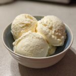 Three generous scoops of pale yellow cottage cheese ice cream served in a small, light blue ceramic bowl.