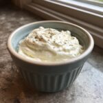 A small, light blue ramekin filled with creamy horseradish sauce for prime rib, topped with cracked pepper.