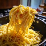 A fork lifts a generous portion of bright, creamy lemon pasta coated in a rich sauce from a skillet.