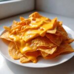 A close-up of tortilla chips smothered in thick, bright orange nacho cheese sauce on a white plate.