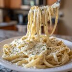 Fork lifting fettuccine noodles coated in creamy olive garden alfredo sauce, topped with Parmesan cheese.