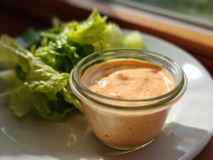 Close-up of a creamy orange-tinted salad dressing in a small glass jar next to fresh lettuce leaves.