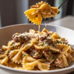 A fork lifts a piece of creamy parmesan garlic beef bowtie pasta, showing the rich sauce dripping back into the bowl.
