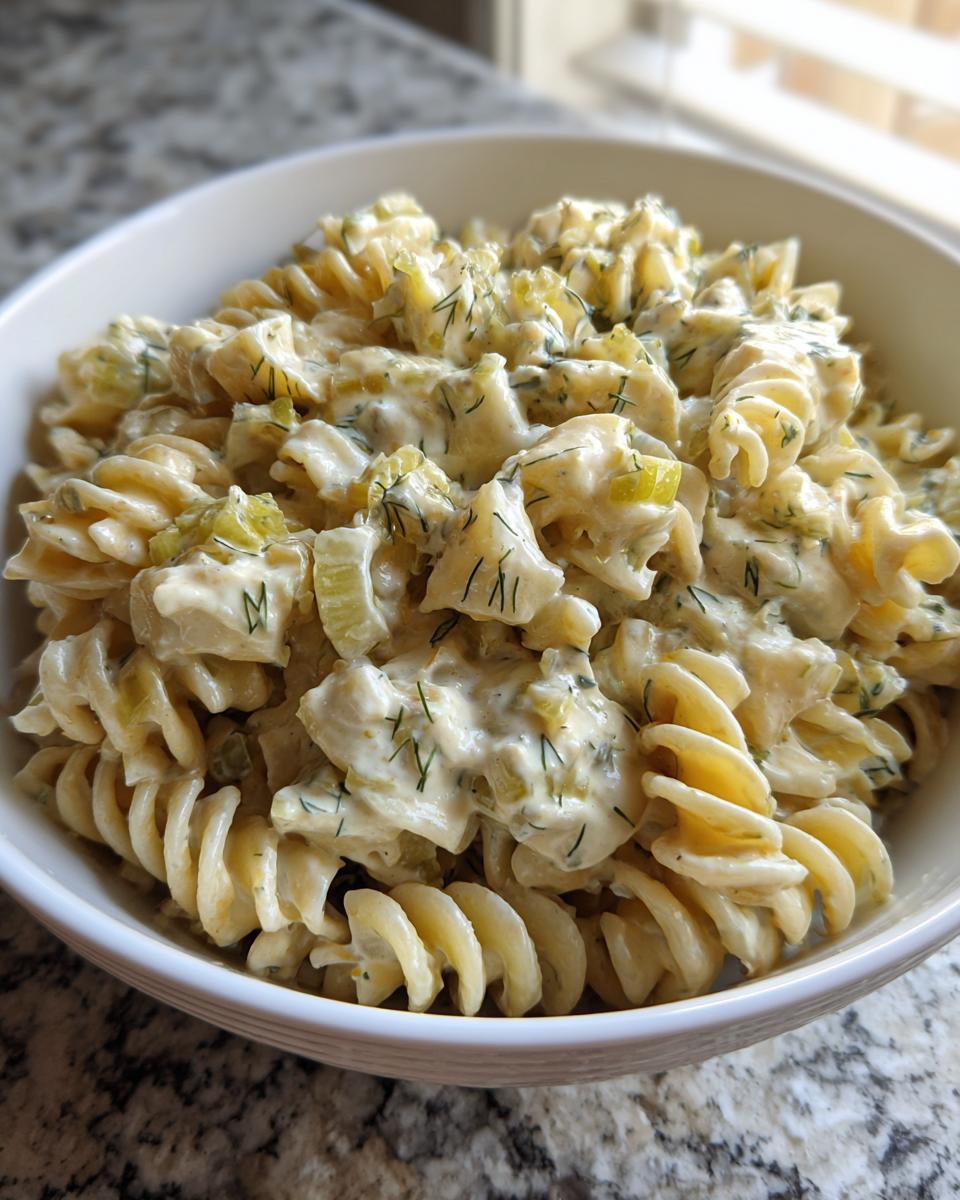 Close-up of creamy pickle pasta salad made with rotini pasta, celery, and fresh dill, served in a white bowl.