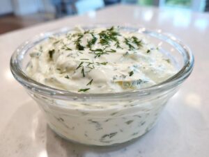 Close-up of creamy, white homemade tartar sauce topped with fresh dill in a clear glass bowl.