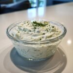 A close-up of creamy homemade tartar sauce, flecked with green herbs and garnished with dill, in a clear glass bowl.